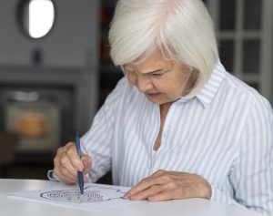 Obrazek przedstawia starszą kobietę (w domyśle z chorobą neurodegeneracyjną), która próbuje rozwiązać trzymanym w ręce długopisem łamigłówkę na zasadzie labiryntu w kształcie ludzkiej głowy/mózgu.