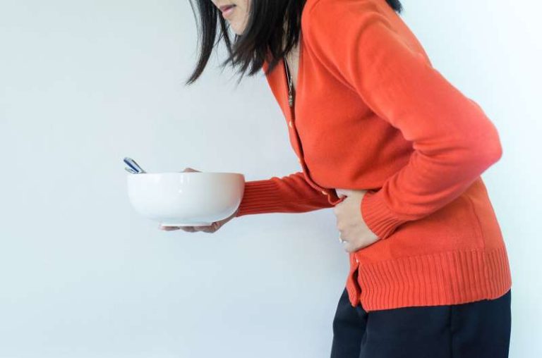 Kobieta na białym tle pochylona do przodu z bólem brzucha. W dłoni trzyma miskę. Drugą dłonią trzyma się za brzuch