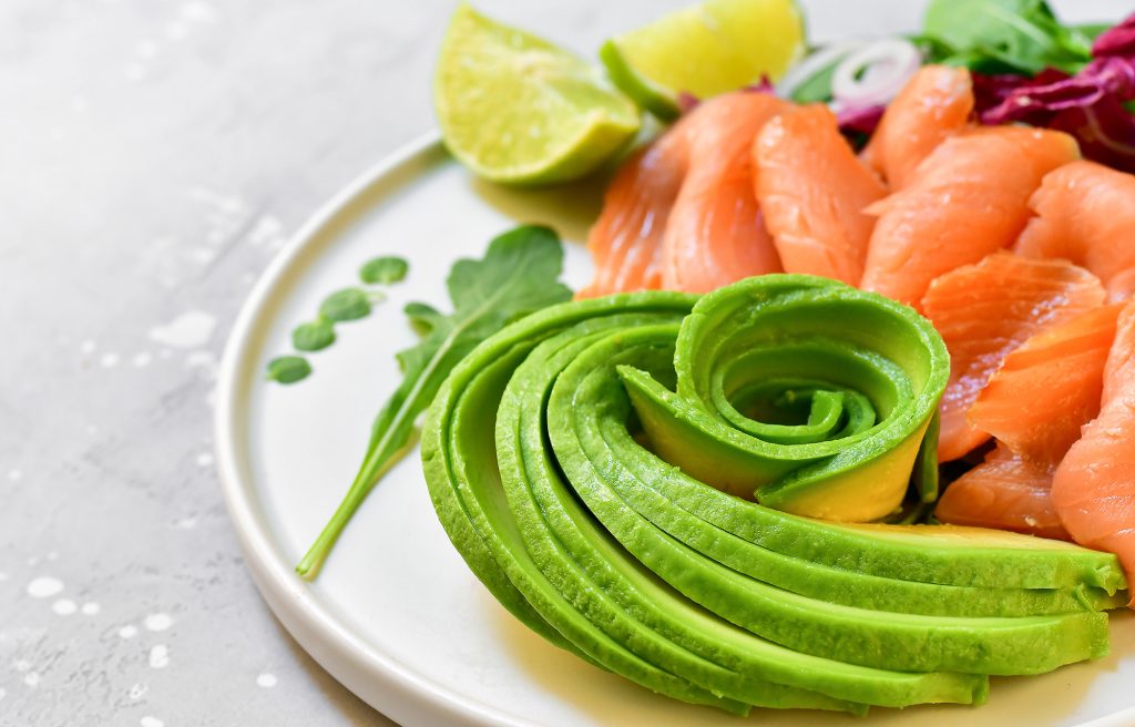 Zdjęcie przedstawia danie na talerzu składające się z pokrojonego awokado i łososia wędzonego, w tle kawałki limonki. Całość w jasnym kolorze.