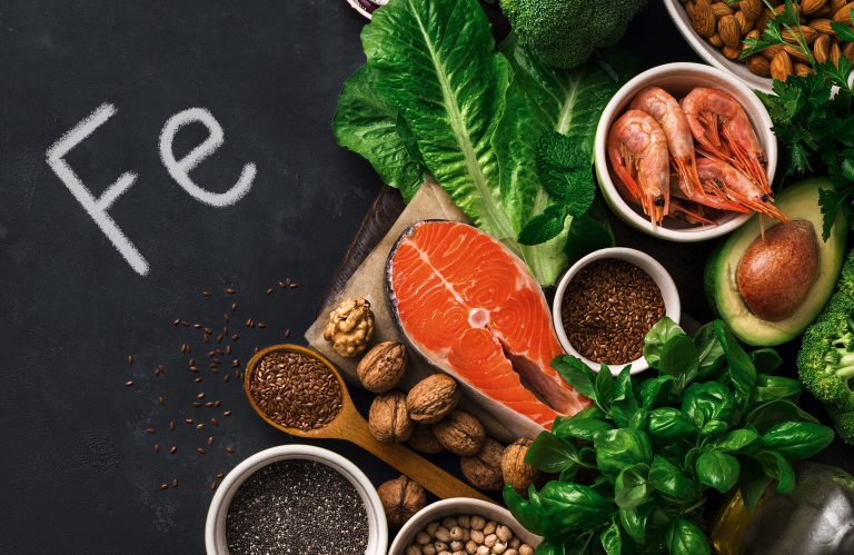 Zdjęcie produktów na czarnym tle, napis Fe czyli żelazo, z prawej strony produkty bogate w żelazo, w tym łosoś, orzechy, nasiona.