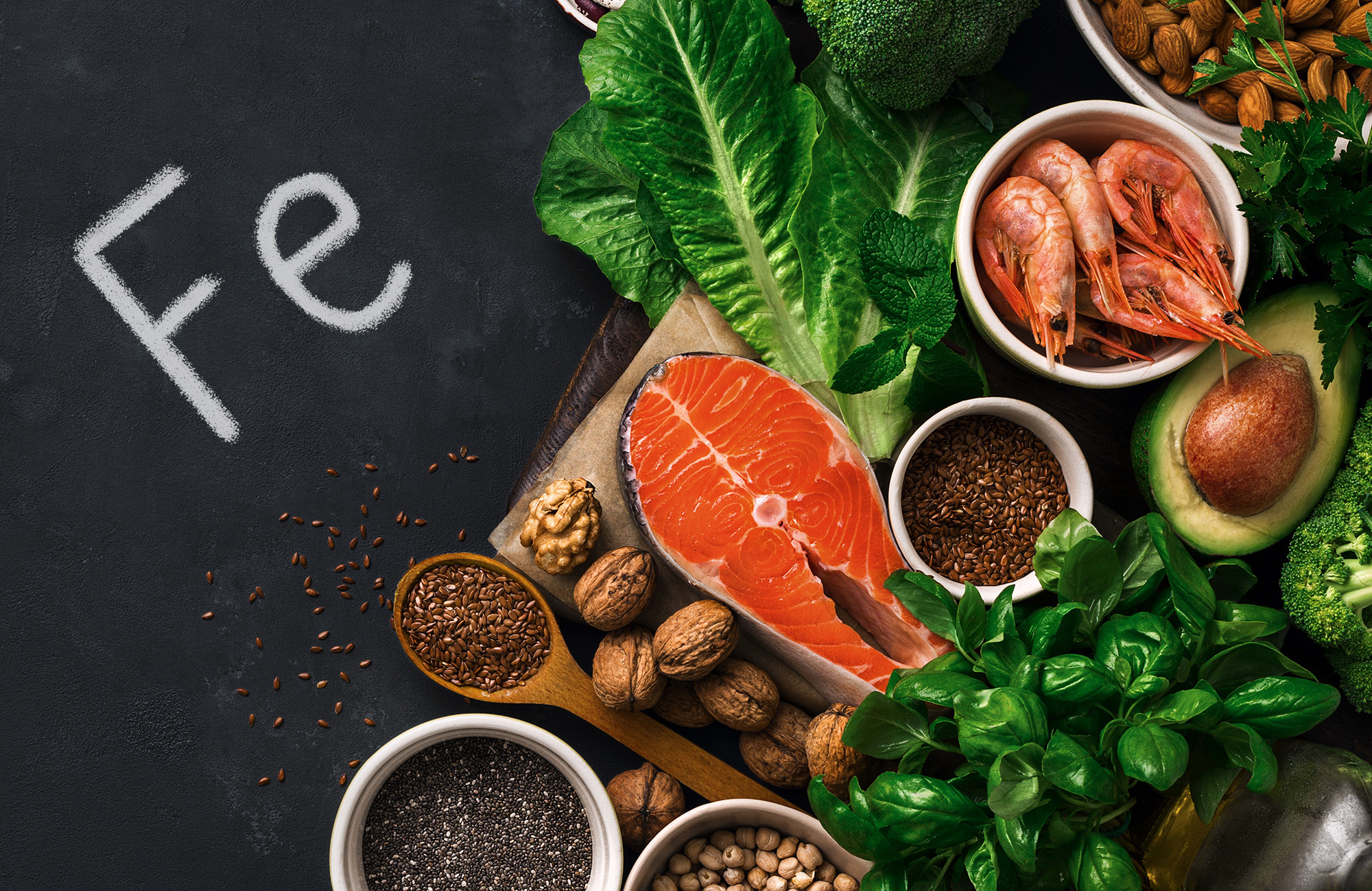 Zdjęcie produktów na czarnym tle, napis Fe czyli żelazo, z prawej strony produkty bogate w żelazo, w tym łosoś, orzechy, nasiona.