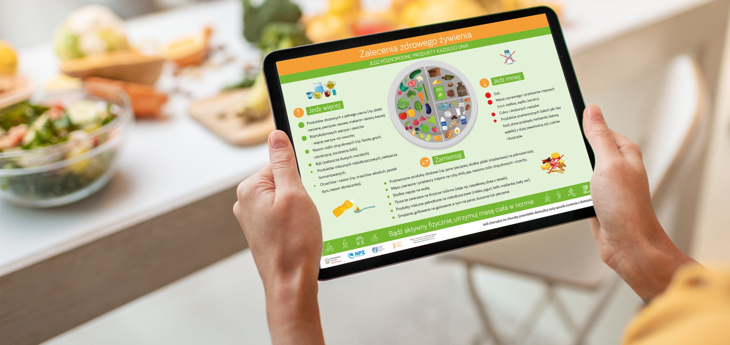 Kobiece ręce trzymające tablet z wyświetlającą się na nim infografiką przedstawiającą Talerz Zdrowego Żywienia, w tle produkty spożywcze w kuchni